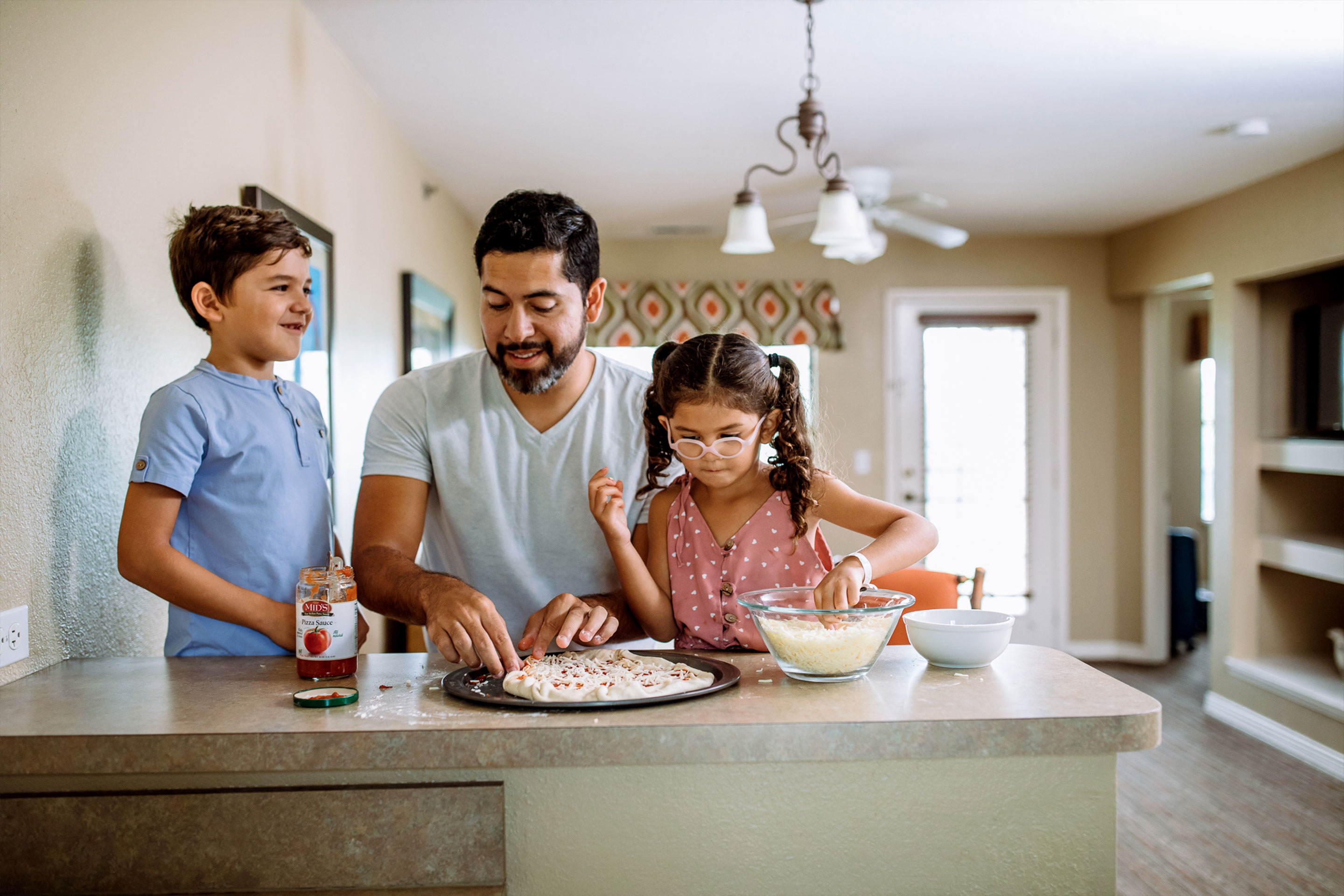 A man (middle), a young boy (left) and girl (right) prepare a pizza at a countertop in a villa.