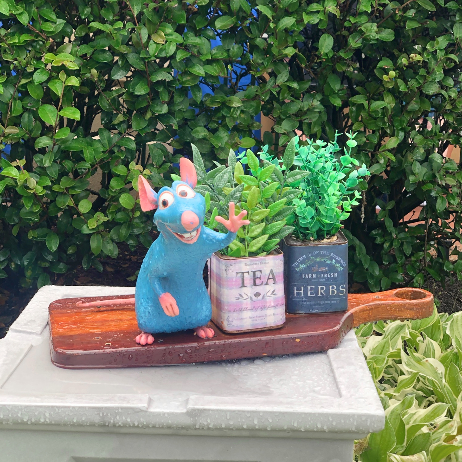 A Remy figurine stands next to canisters that read, 'Tea Leaves' and 'Herbs' near shrubbery.