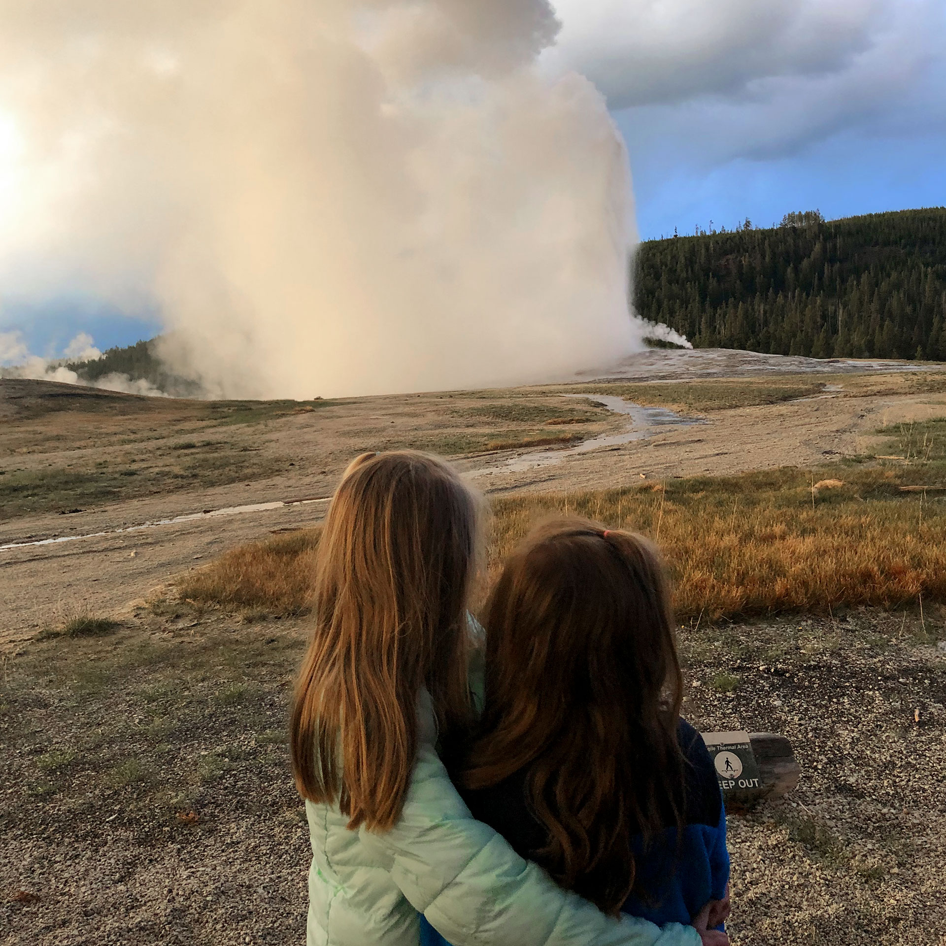 Author, Chris Johnstons' daughters, Kyndall (left), and Kyler (right) face out to watch Old Faithful at dusk while wrapping one arm around each other.