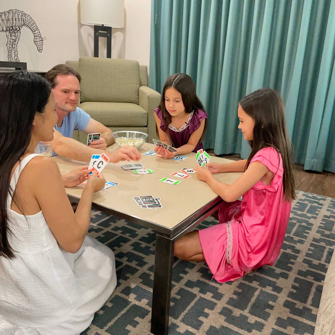 (From left to right) A woman, man and two young girls play a card game while kneeling near a coffee table and green chair indoors.