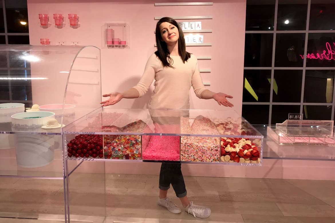 A woman in white sweater, blue jeans and white sneakers stands in a pink room surrounded by ice cream counter with toppings.