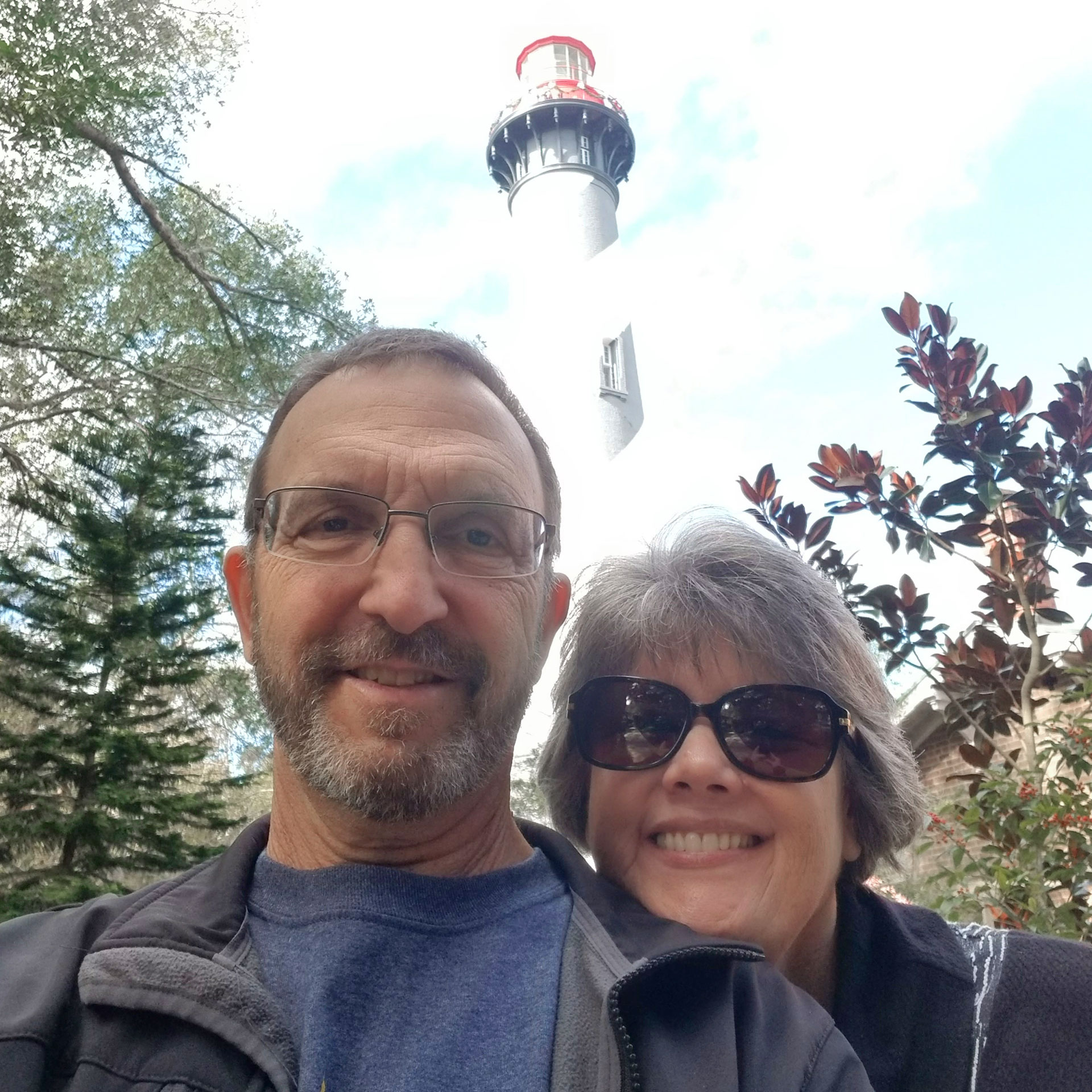 A woman in sunglasses and a jacket stands behind a man in reading glasses and a grey jacket near a white lighthouse.