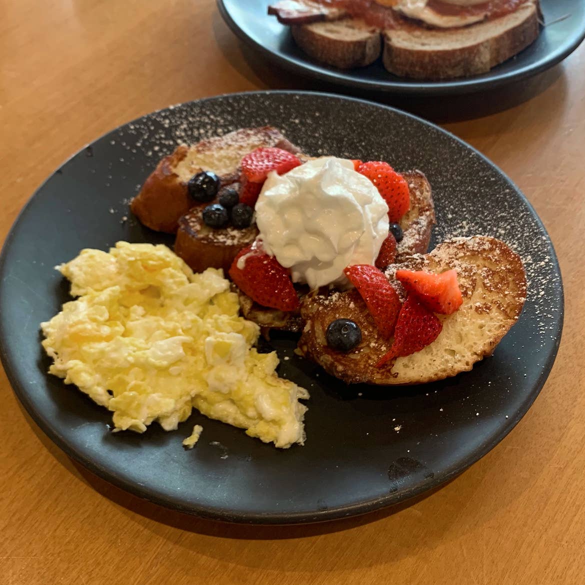 A black plate on a wooden dining table contains scrambled eggs, three french toast slices covered with whipped cream, berries and powdered sugar.