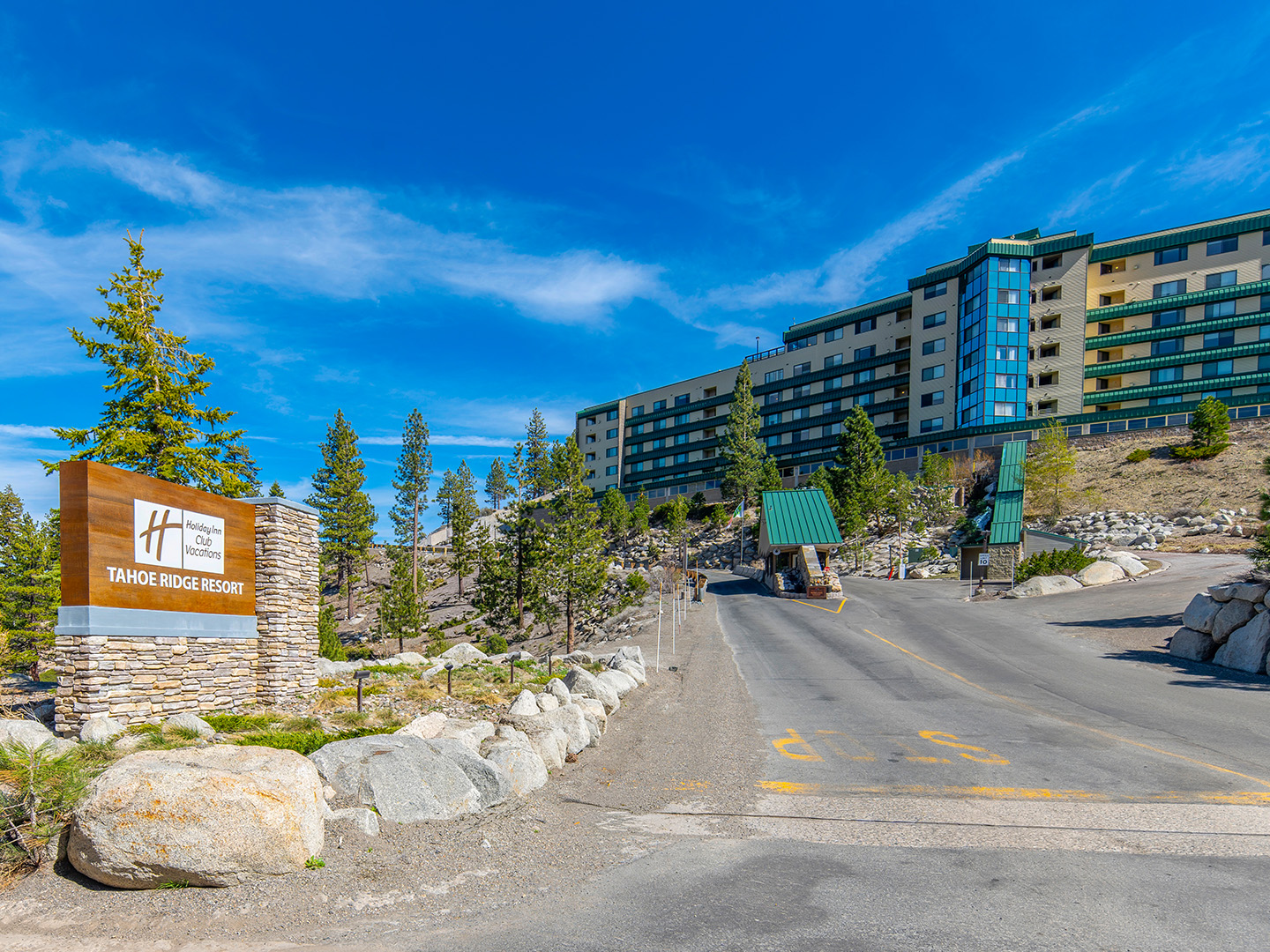 Entrance to Holiday Inn Club Vacations Tahoe Ridge Resort featuring a stone monument sign, uphill driveway with guard gate, and a multi-story resort building set among pine trees under a blue sky.