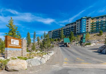 Entrance to Holiday Inn Club Vacations Tahoe Ridge Resort featuring a stone monument sign, uphill driveway with guard gate, and a multi-story resort building set among pine trees under a blue sky.