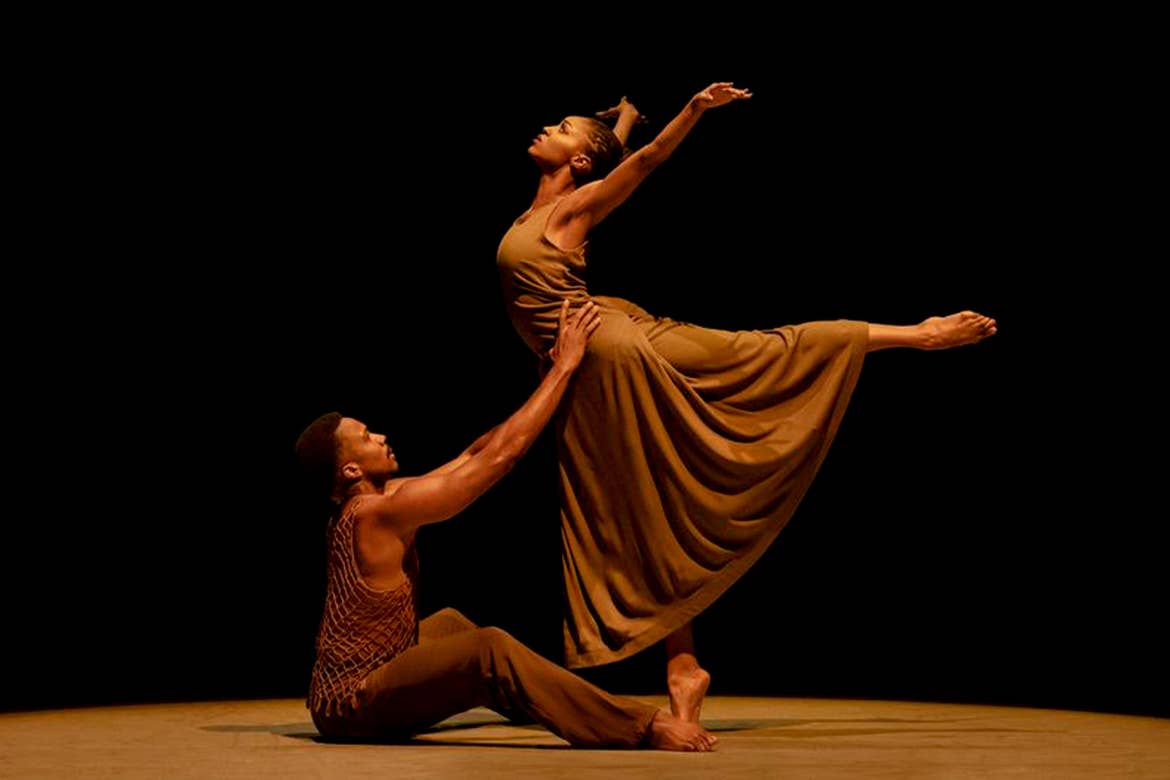 A woman and male performer wearing brown dancewear are dynamically posing on-stage under a spotlight.