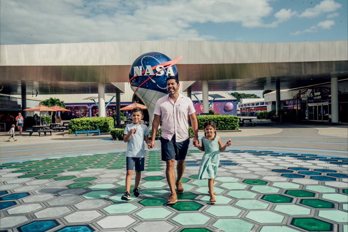 A man wearing a pink button-up and grey shorts holds the hands of a young boy in a blue shirt and grey shorts and girl in blue dress as they walk in front of a NASA globe outdoors.