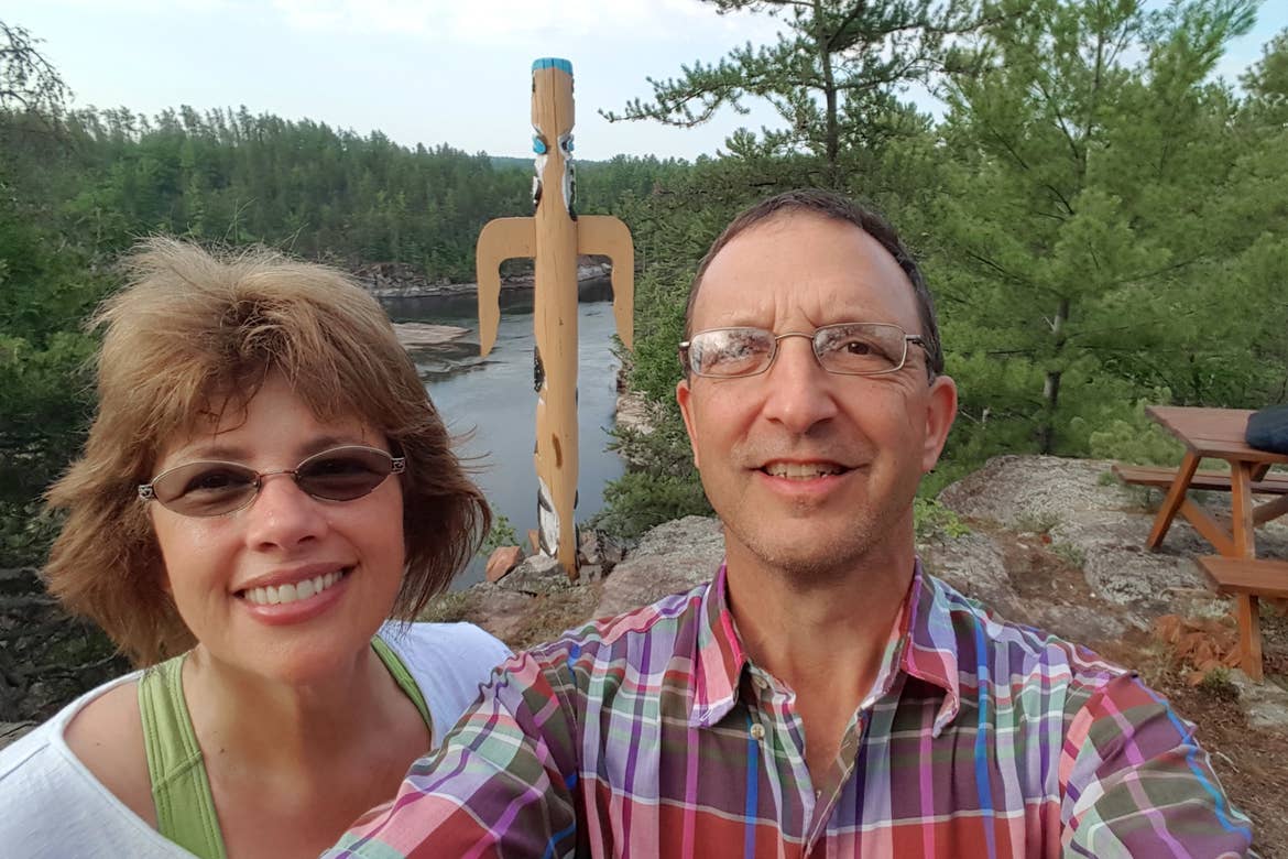 a caucasian woman wearing a green camisole under a white t-shirt (left)n and a caucasian man wearing a plaid button-up shirt (right) stand near a totem at the Dokis First Nation Reservation in Ontario, Canada.