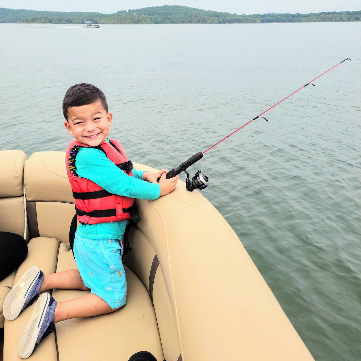 Angelica's son goes fishing off the side of a tritoon wearing a life jacket and aqua swimwear.