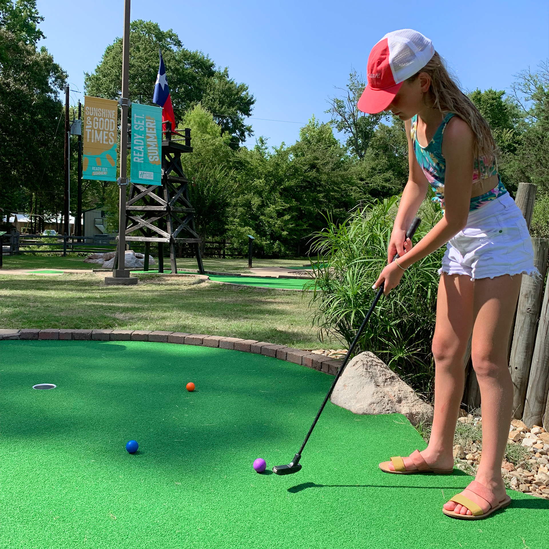 A caucasian girl stands with her putter on a mini golf course.