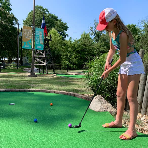 A caucasian girl stands with her putter on a mini golf course.