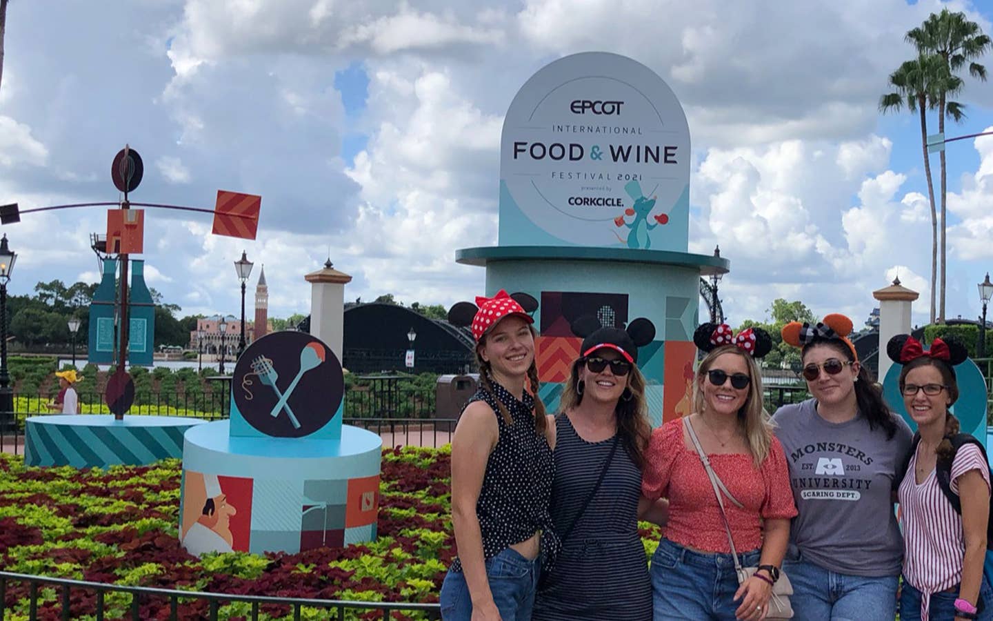 Five caucasian women wearing Mickey and Minnie ears stand in front of a display for the Epcot International Food & Wine Festival near the World Showcase Lagoon.