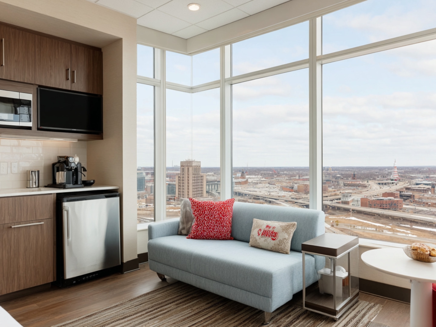 Spacious corner hotel suite with expansive windows, comfortable sofa, and fully equipped kitchenette.