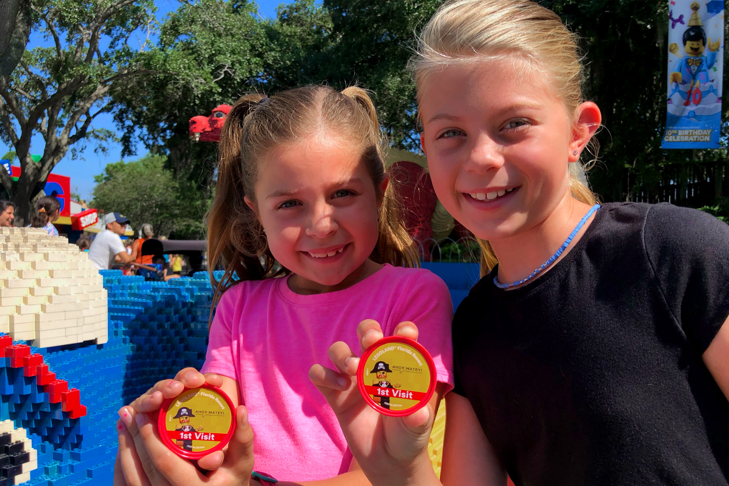 A young girl in a pink t-shirt (left) and another in a black t-shirt (right) stands next to an oversized LEGO figure holding pins.