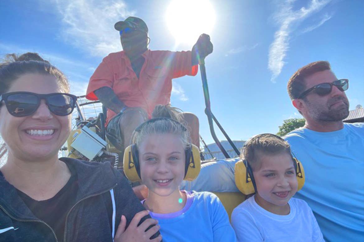 A caucasian woman (left) wearing sunglasses and a black zip-up hoodie, two Caucasian girls (middle) and a caucasian male (right) wearing sunglasses and a blue long-sleeve shirt sit on an airboat in the Everglades.