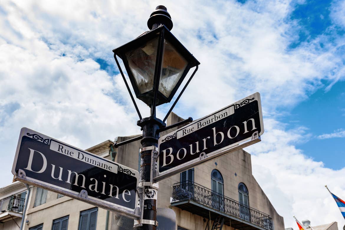 A black lamppost indicating the streets of 'Dumaine' (left) and 'Bourbon' (right) under a cloudy blue sky.
