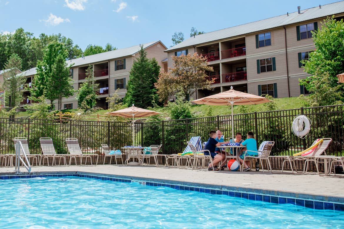 A caucasian family of five (left to right: Two tween boys, a young girl, a woman and man) sit at a table under an umbrella near an outdoor pool surrounded by green trees and the exteriors of our Oak and Spruce resort located in Lee, Massachusetts.