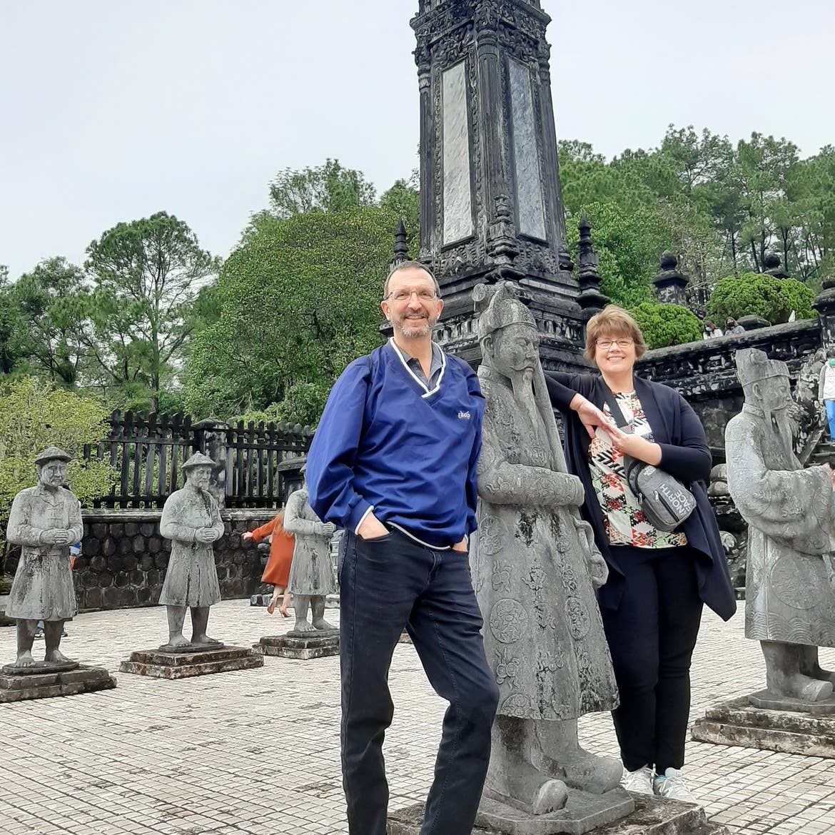 A caucasian male (left) wearing a navy windbreaker and jeans and a caucasian woman wearing a black tracksuit and a travel bag stand at a temple in Da Nang Vietnam surrounded by statues.