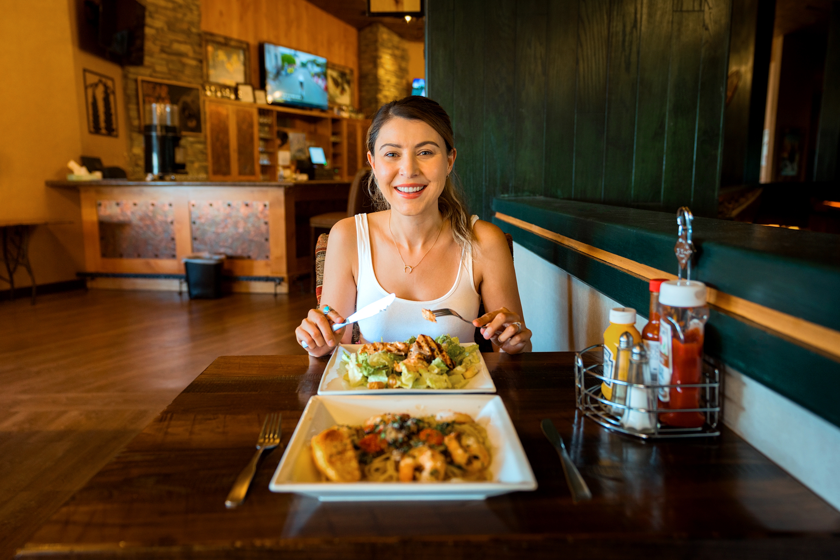 A caucasian woman wearing a white tank top sits at a wooden table with a knife and fork as two white, square plates of food sit on the table indoors.