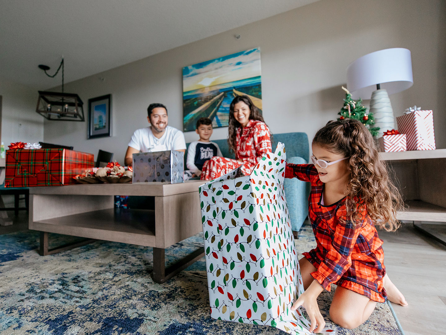 A man, young boy and woman watch a young girl unwrap a present in a living room while all wearing pajamas.