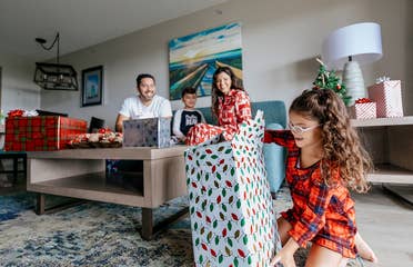 A man, young boy and woman watch a young girl unwrap a present in a living room while all wearing pajamas.