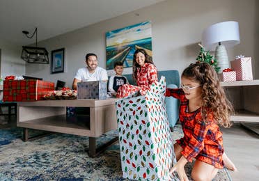 A man, young boy and woman watch a young girl unwrap a present in a living room while all wearing pajamas.