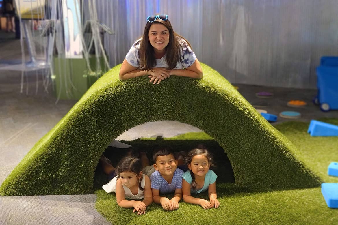A woman lays on a mossy bridge where three toddlers sit below in an indoor space.