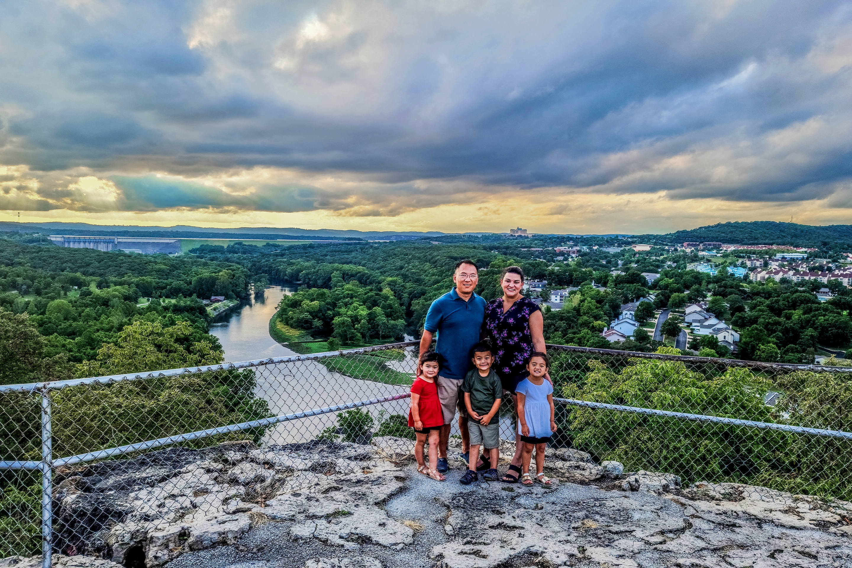 Three toddlers (Two girls (front-left and right) and a boy) stand in front of a man (back-left) and woman (back-right) near a chain link fence overlooking the river in Branson, Missouri.