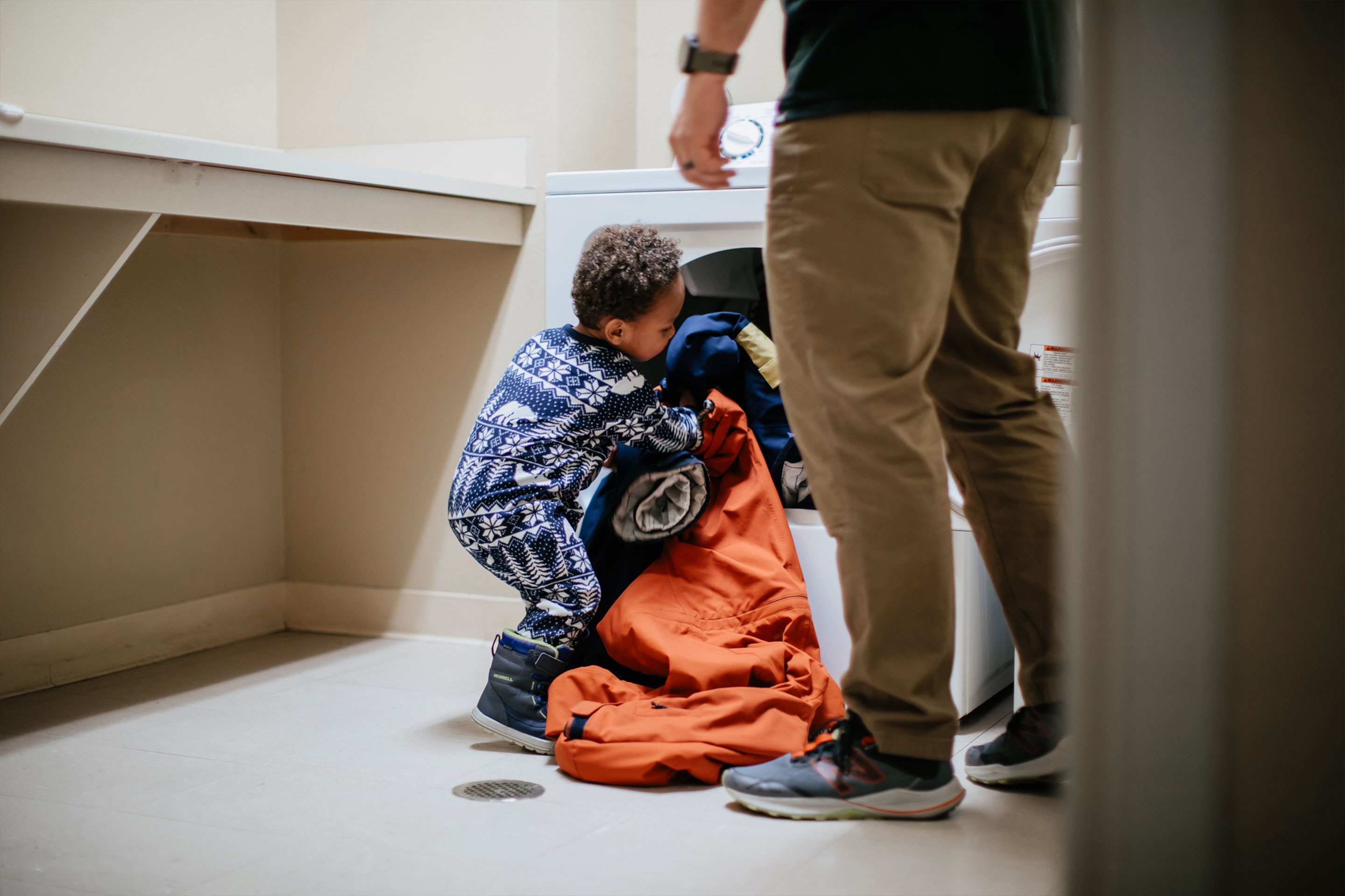 A boy toddler loads laundry into a front-loading washing machine as a man stands nearby.