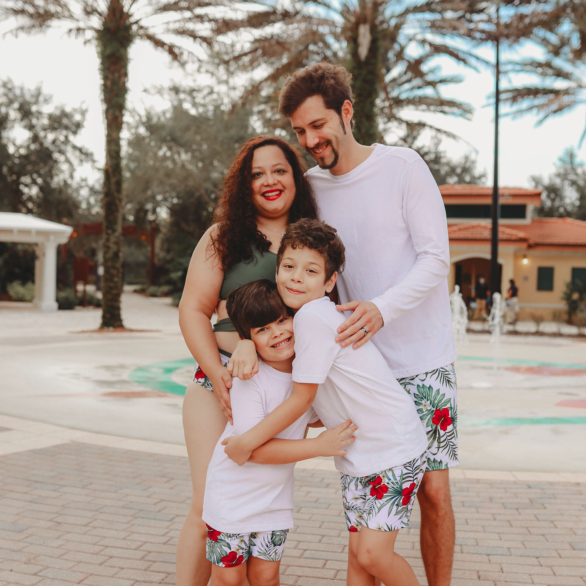 A woman, man and two young boys wear swimwear while standing near an outdoor splash pad.