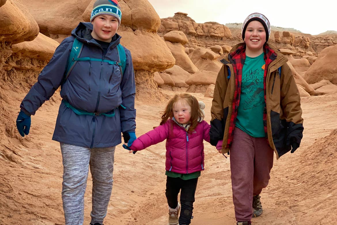 Featured Contributor, Melody Forsyth's two sons (left and right) walk with daughter, Ruby (middle) in front of a rock formation holding hands while wearing hiking backpacks.