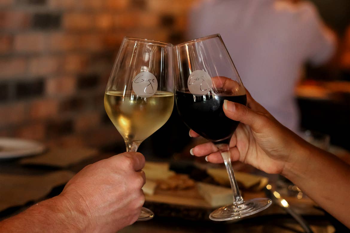 A pair of hands hold two glasses of white wine (left) and red wine (right) inside of a restaurant.