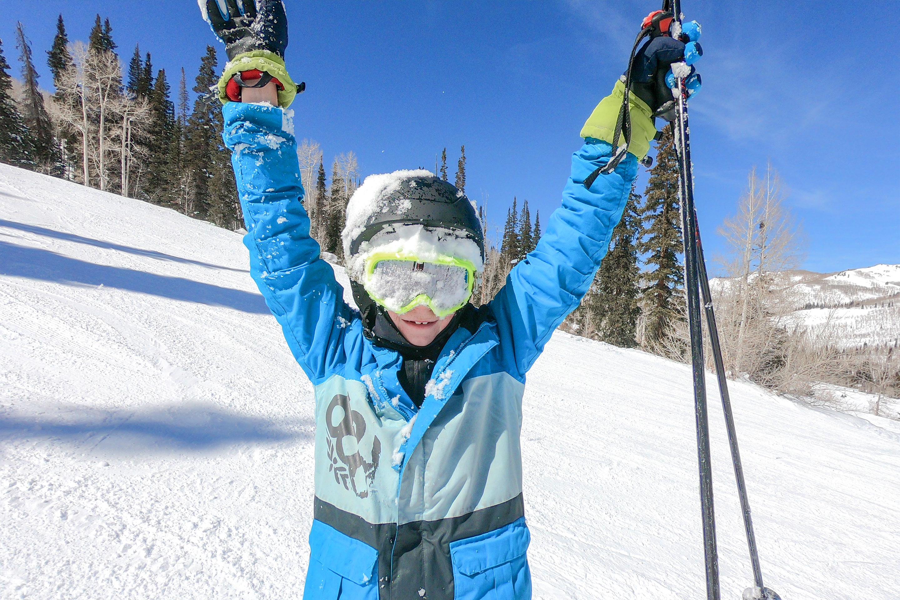 A young boy wears a black ski helmet, neon yellow goggles and winter jacket, and holding a set of ski poles while standing outdoors in the snow.