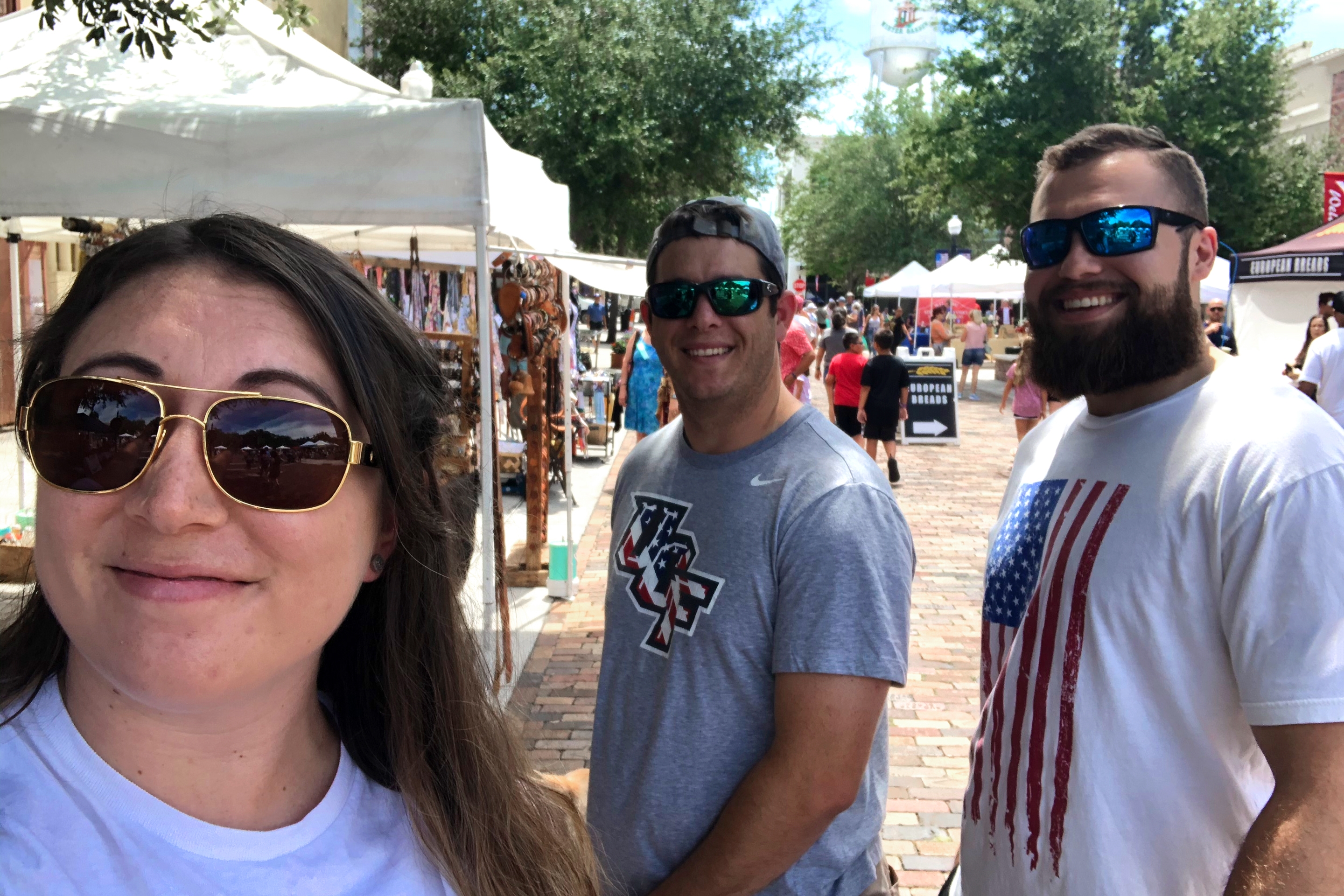 A woman (left) wearing a white t-shirt and sunglasses stands in front of two men (right) wearing t-shirts and sunglasses in front of the Winter Garden Famers Market.