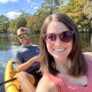 A woman wearing a coral tank top and sunglasses sits in a kayak with a man in a baseball cap, grey shirt and dark shorts on open water.