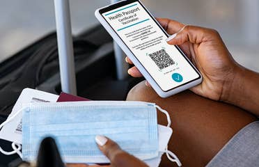Traveler pulling out their cell phone to look at their digital health pass/vaccine passport, with a mask in their hand and luggage in front of them.