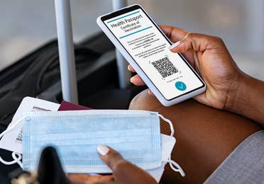Traveler pulling out their cell phone to look at their digital health pass/vaccine passport, with a mask in their hand and luggage in front of them.