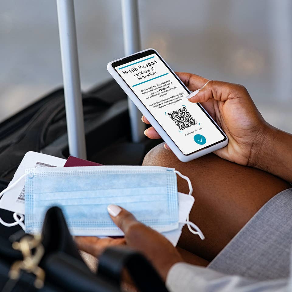 Traveler pulling out their cell phone to look at their digital health pass/vaccine passport, with a mask in their hand and luggage in front of them.