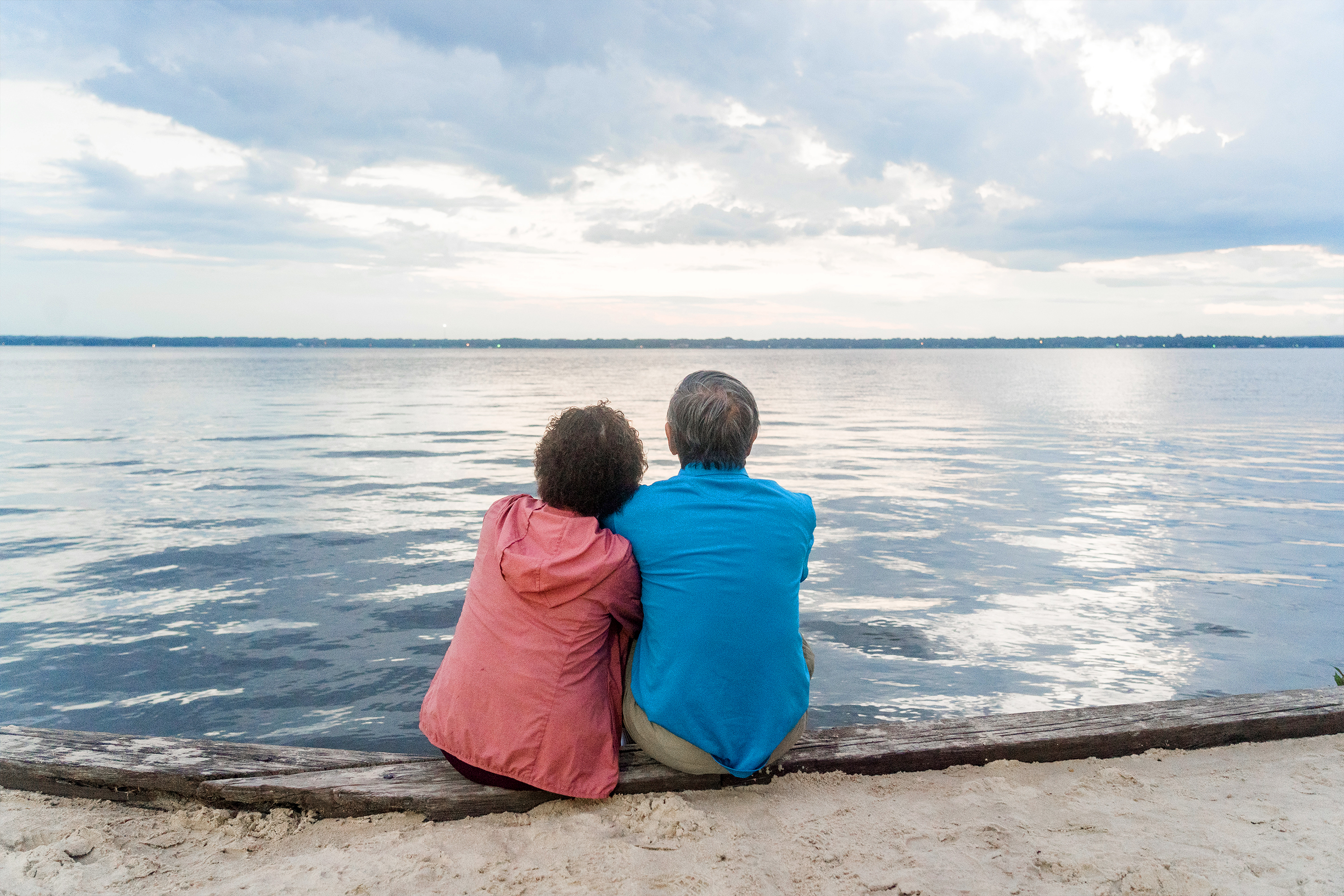 An older Asian woman (left) wears a pink zip-up hoodie next to an older Asian man (right) wearing a blue polo and khaki shorts as they sit on a beach dipping their toes into the water.