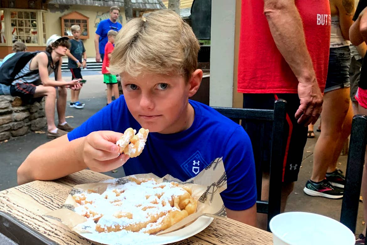 A boy in a blue t-shirt sits at a wooden dining table eating funnel cake with powdered sugar.