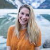 Featured Contributor, Ashlyn George, wears an orange t-shirt in front of an icy river and snowcapped mountains.
