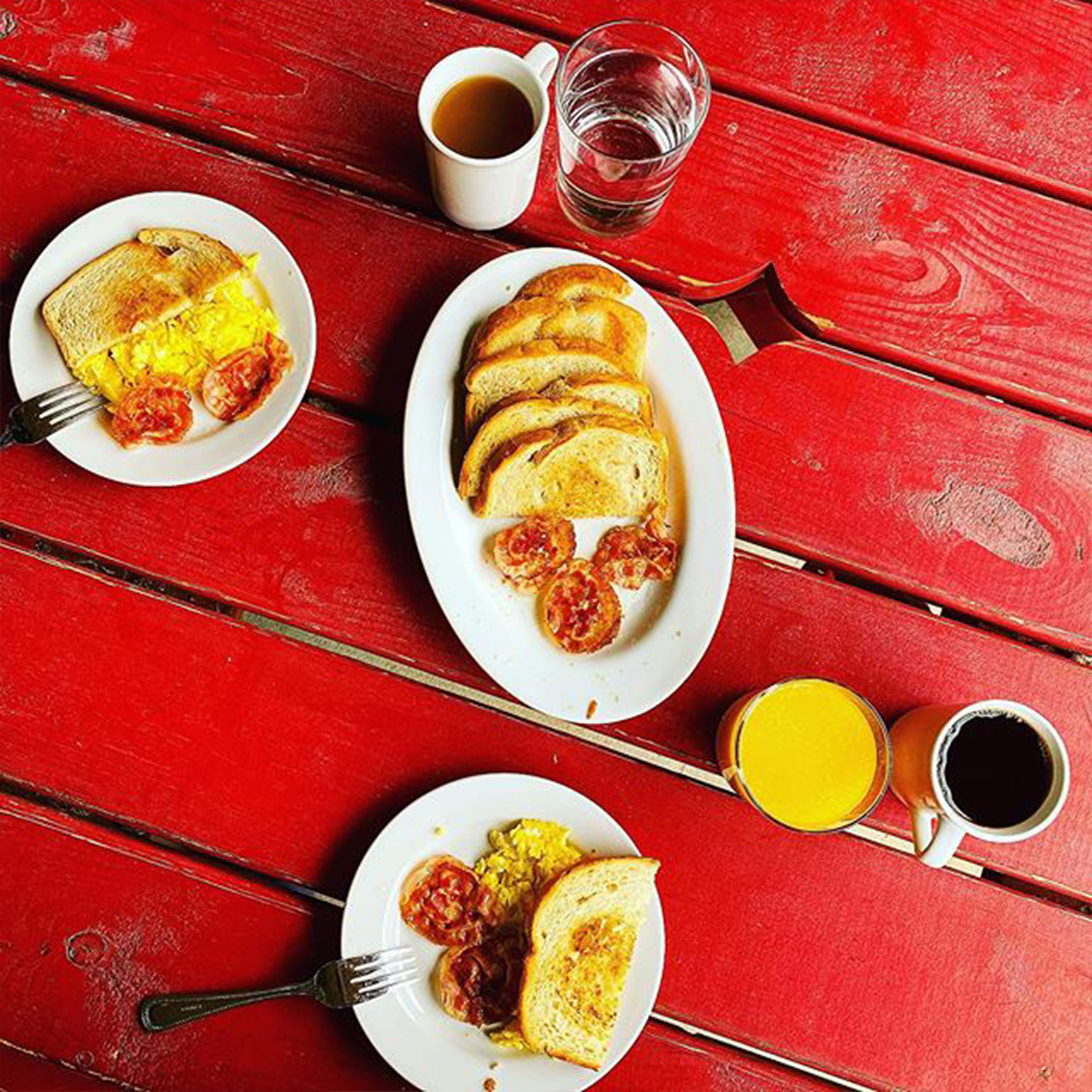 Three plates of egg and toast with beverages are placed on a red picnic table.