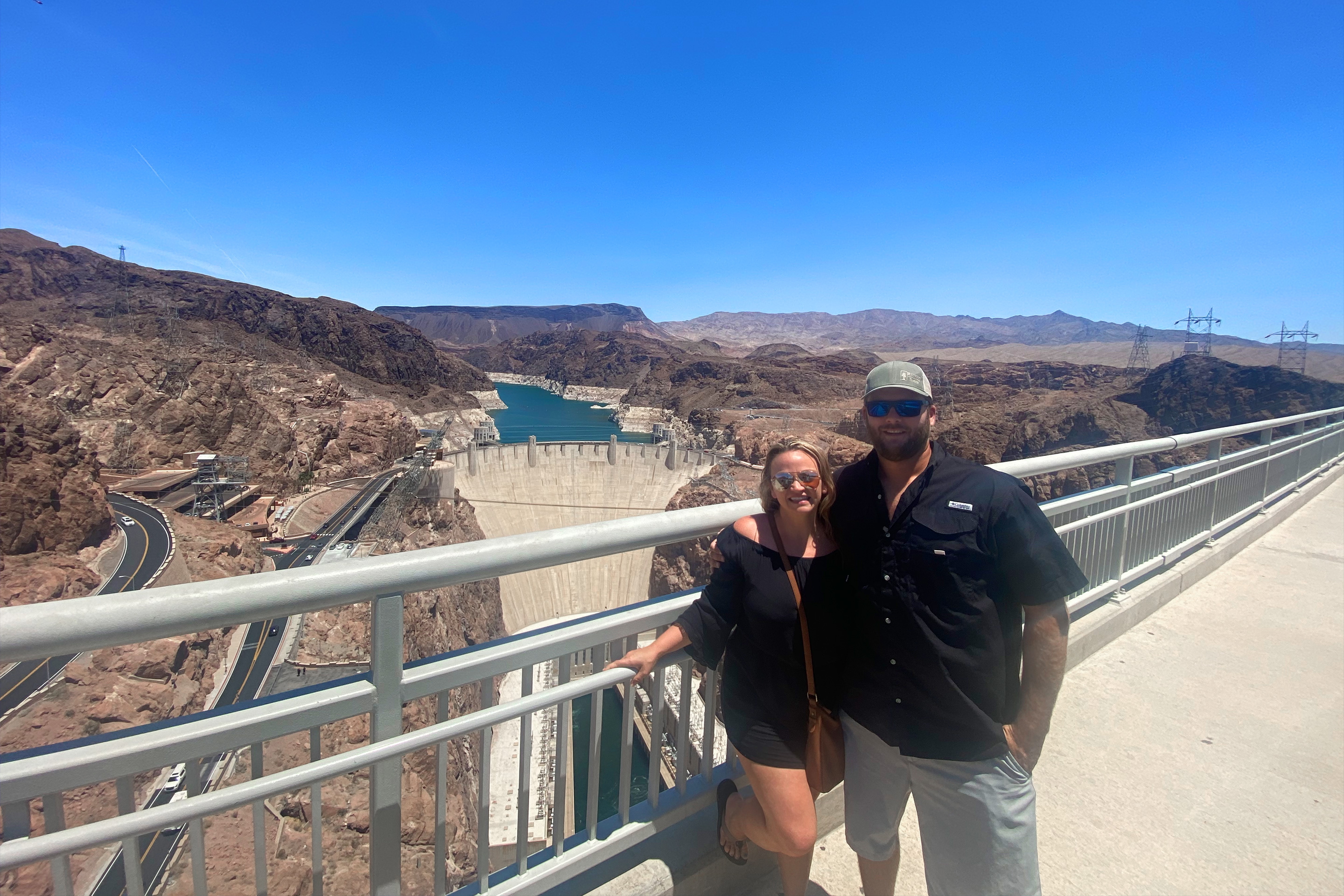 A woman wearing sunglasses (left) poses near a man wearing sunglasses and a hat (right) in front of the Hoover Damn under a clear blue sky.