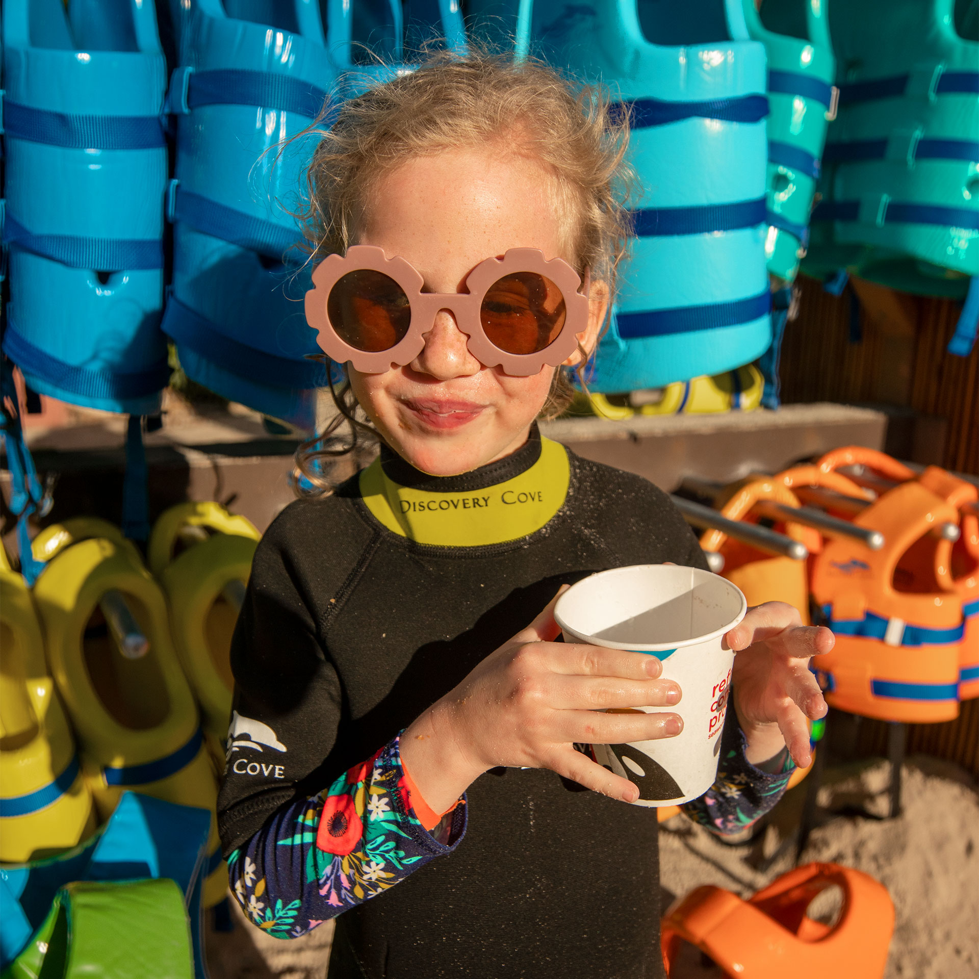 A young girl holds a white cup wearing pink, floral sunglasses and a black and yellow wetsuit near a rack of multi-colored lifejackets.