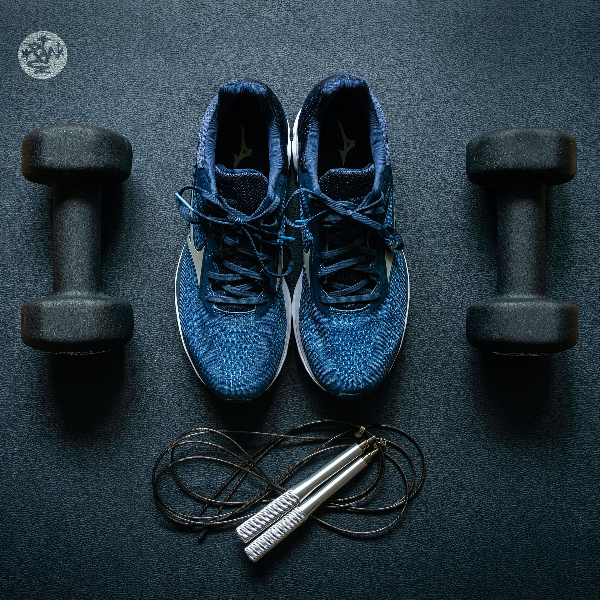 A pair of blue gym shoes, two black free weights, and a jump rope lay out over a navy yoga mat.