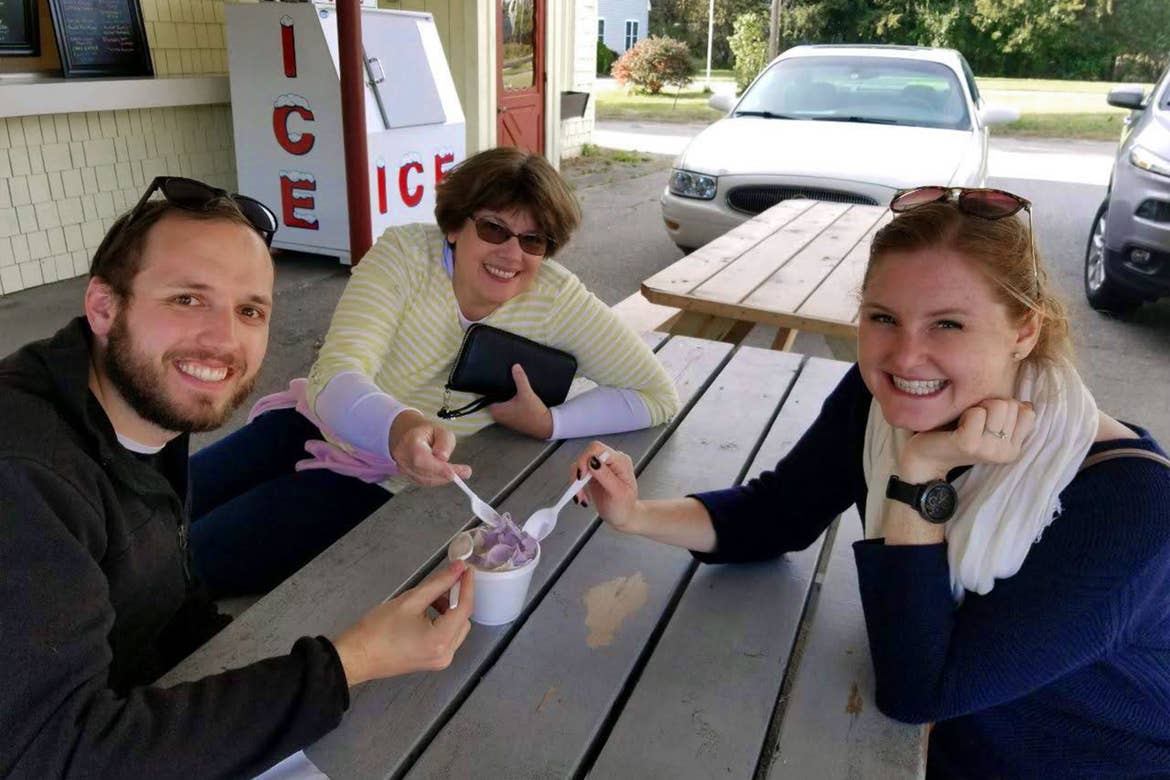 From left to right: A man, a woman and another woman sit at a picnic table sharing some ice cream on a covered outdoor patio.