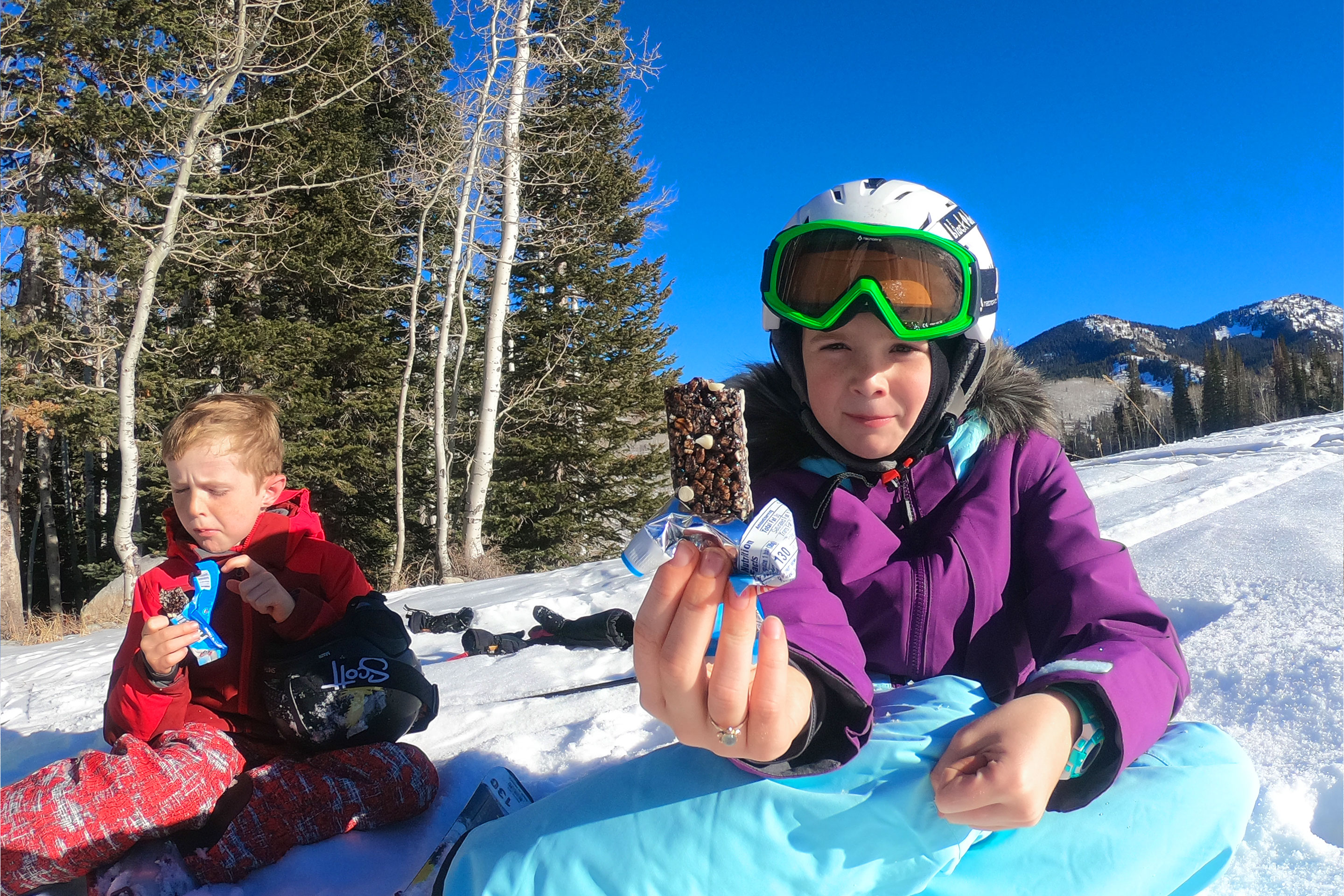 Two young children wearing winter and skiing gear sit on a snowy slope enjoying treats.