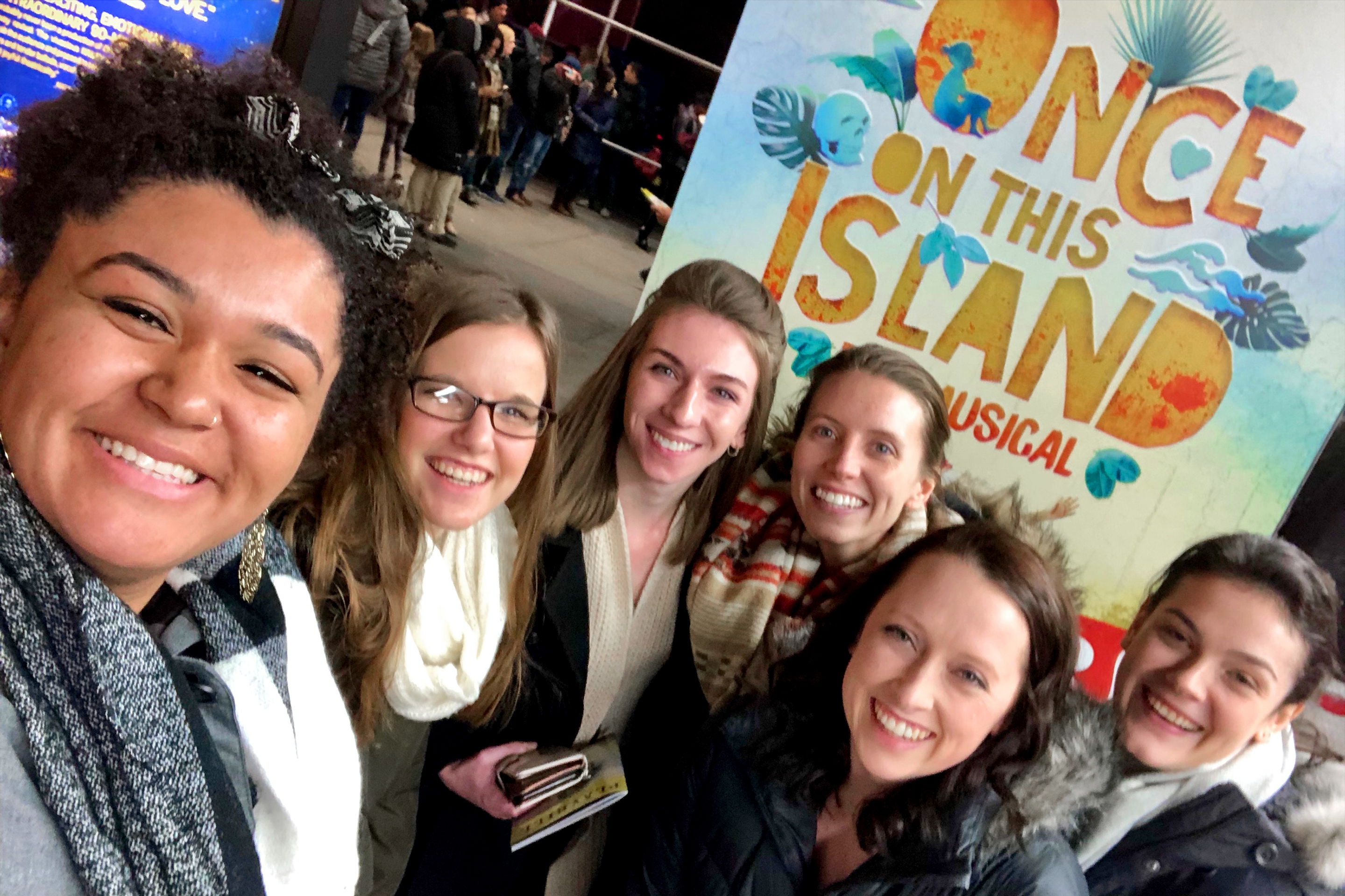 Several women stand wearing scarves and winter coats in front of a sign that reads, 'Once On This Island - A Musical' in New York City.