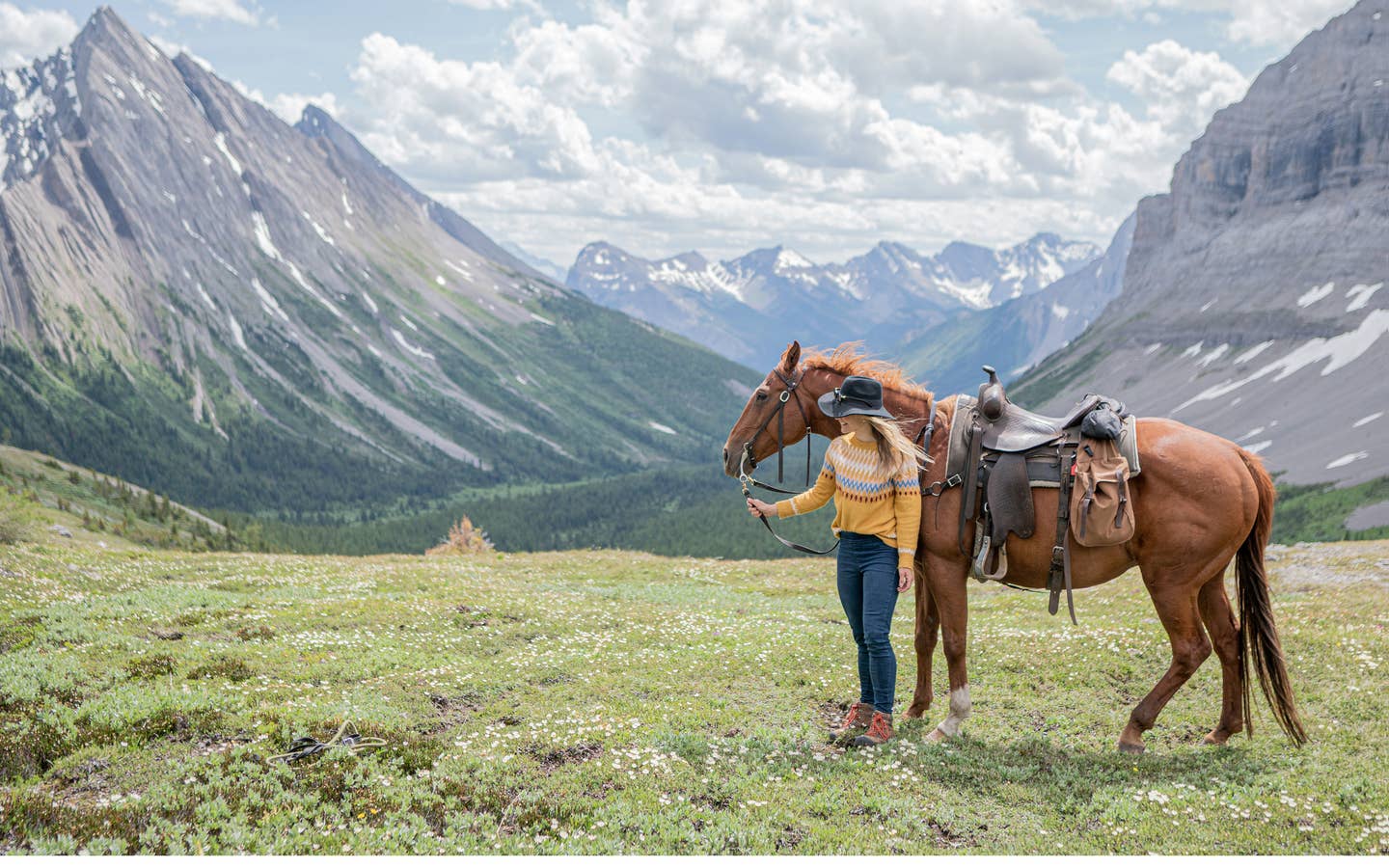 Featured Contributor, Ashlyn George, stands with a horse in front of the Canadian mountain range.