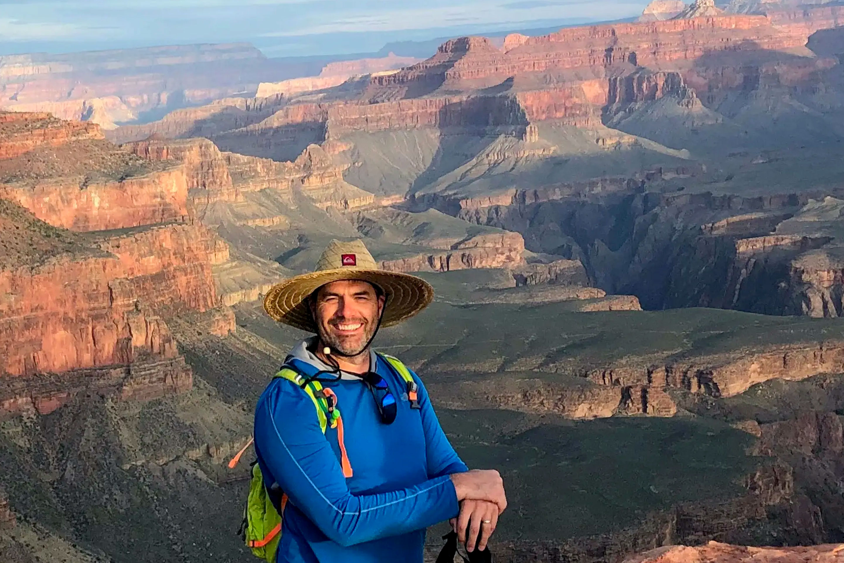 A caucasian male wearing a blue long-sleeve shirt and sunhat stands in front of the beautiful, mountainous Grand Canyon landscape.